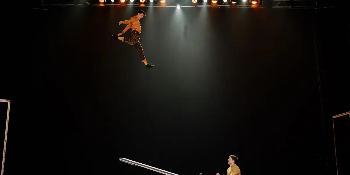 Un interprète est lancé dans les airs depuis une bascule de Léonard Meylan et Nathan Sterchi, sur une scène à l'éclairage dramatique et à l'arrière-plan sombre.