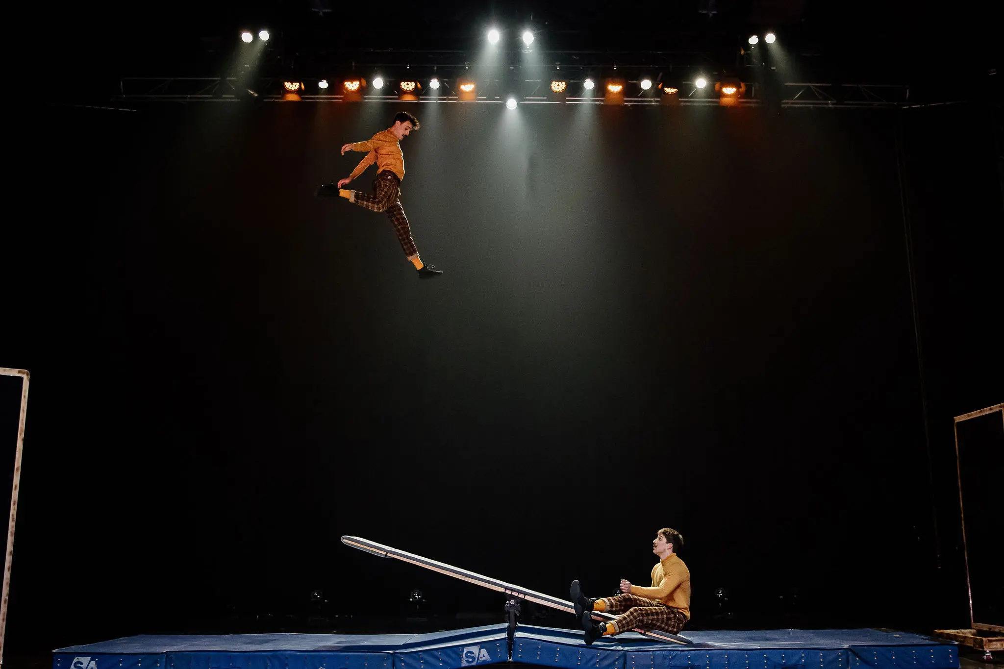 Un interprète est lancé dans les airs depuis une bascule de Léonard Meylan et Nathan Sterchi, sur une scène à l'éclairage dramatique et à l'arrière-plan sombre.