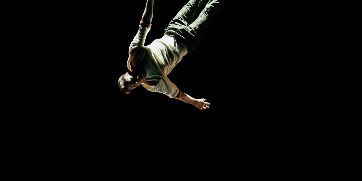 Une personne en tenue décontractée est suspendue à une corde, la tête en bas, sur un fond sombre, éclairée par une seule source lumineuse - une scène qui rappelle le style évocateur de Marceau Bidal.