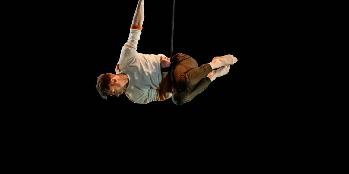 Marceau Bidal, vêtu d'une chemise blanche et d'un pantalon marron, est suspendu dans les airs par des cordes et exécute un numéro aérien sur un fond noir et sombre.