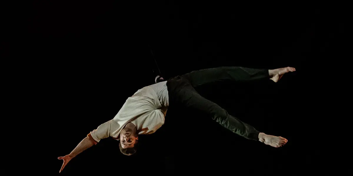 Une personne vêtue d'une chemise blanche et d'un pantalon sombre est suspendue à l'envers dans les airs sur un fond noir, adoptant une pose dynamique rappelant celle de Marceau Bidal, avec un bras et les deux jambes tendus vers l'extérieur.
