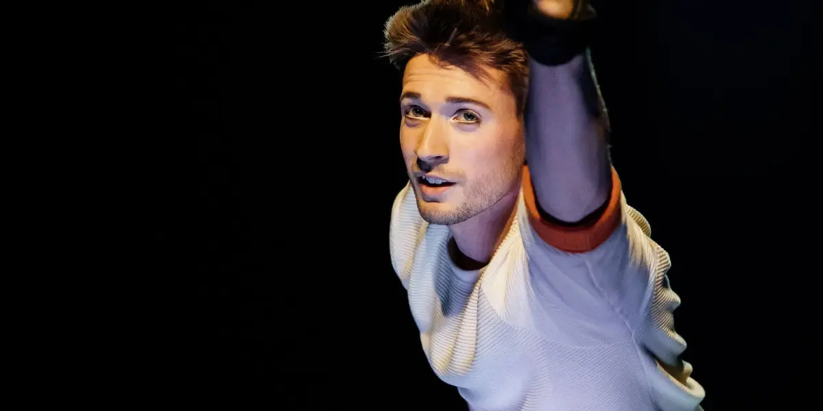 Un homme vêtu d'une chemise blanche et d'un pantalon sombre exécute un numéro aérien, en saisissant des sangles noires et en tendant un bras vers l'avant. Marceau Bidal se détache sur un fond sombre avec un éclairage dramatique.