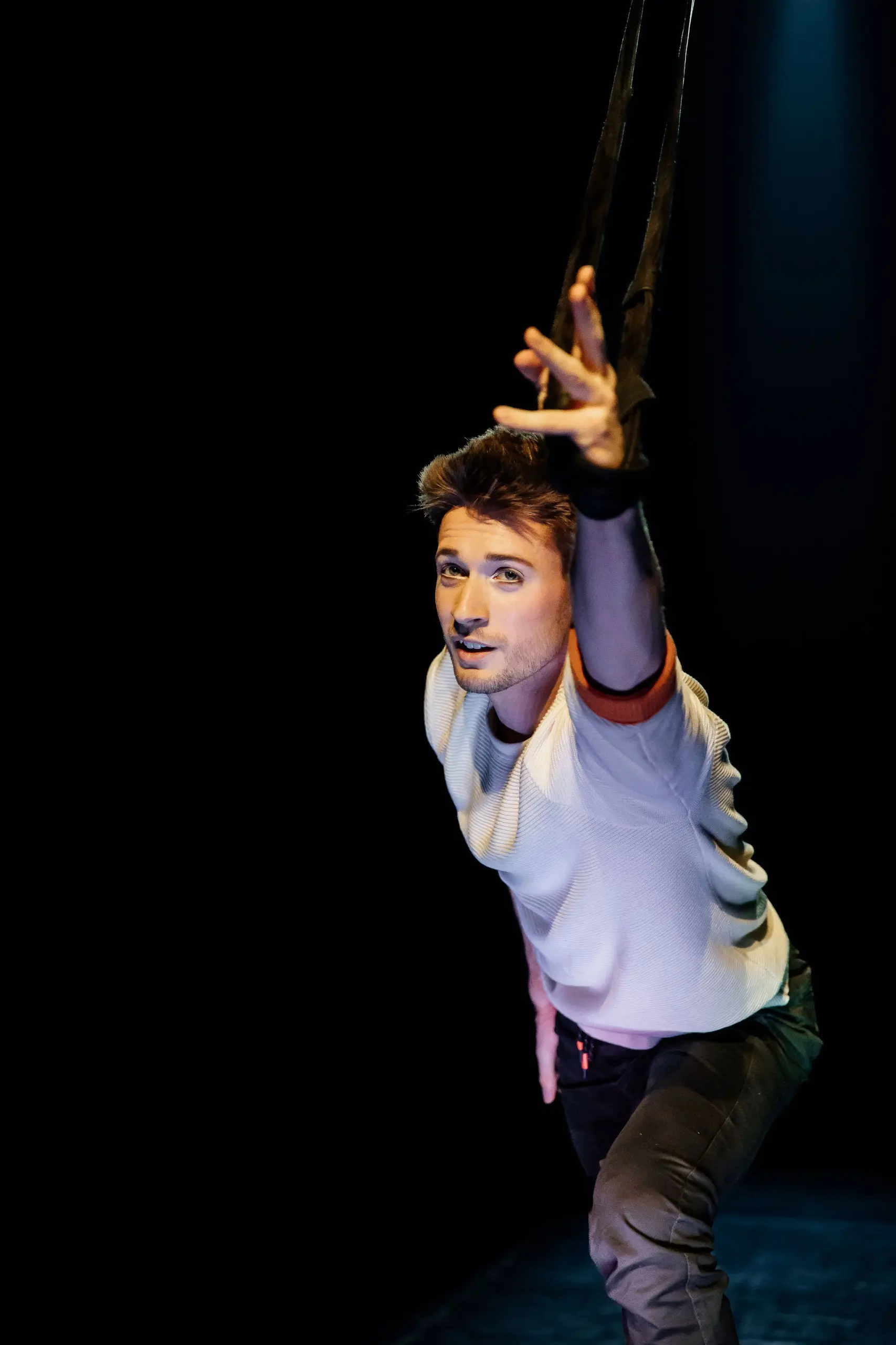 Un homme vêtu d'une chemise blanche et d'un pantalon sombre exécute un numéro aérien, en saisissant des sangles noires et en tendant un bras vers l'avant. Marceau Bidal se détache sur un fond sombre avec un éclairage dramatique.