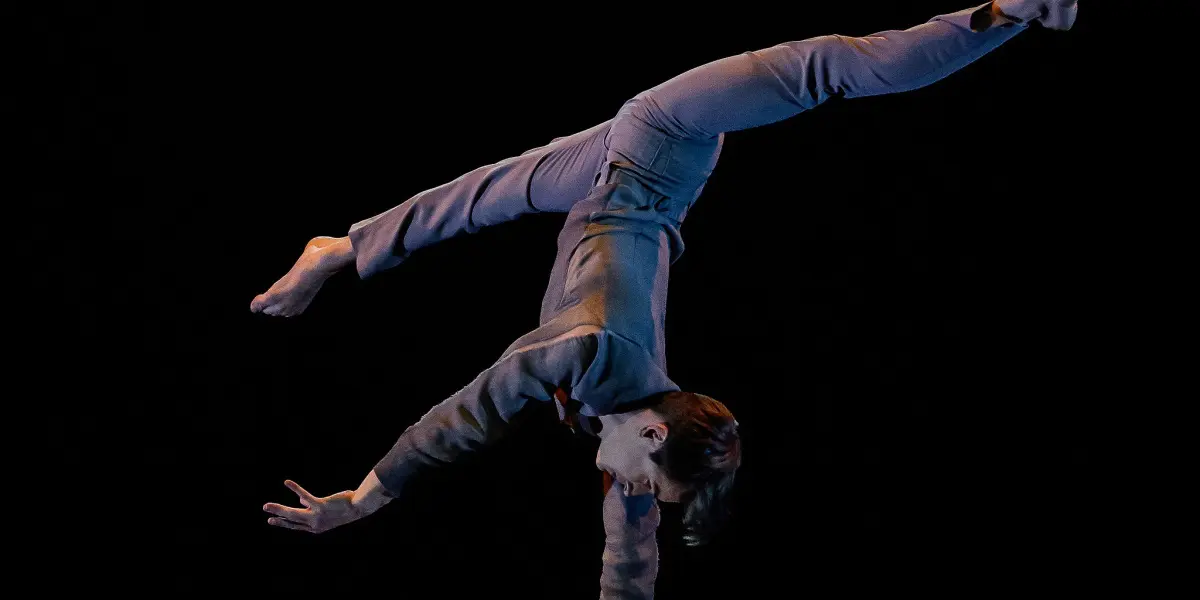 Louis-Thomas Gauthier, vêtu d'une tenue grise, se tient en équilibre sur une main au sommet de quatre grands supports sur un fond sombre, les jambes tendues en fente, faisant preuve d'une force et d'une souplesse remarquables.