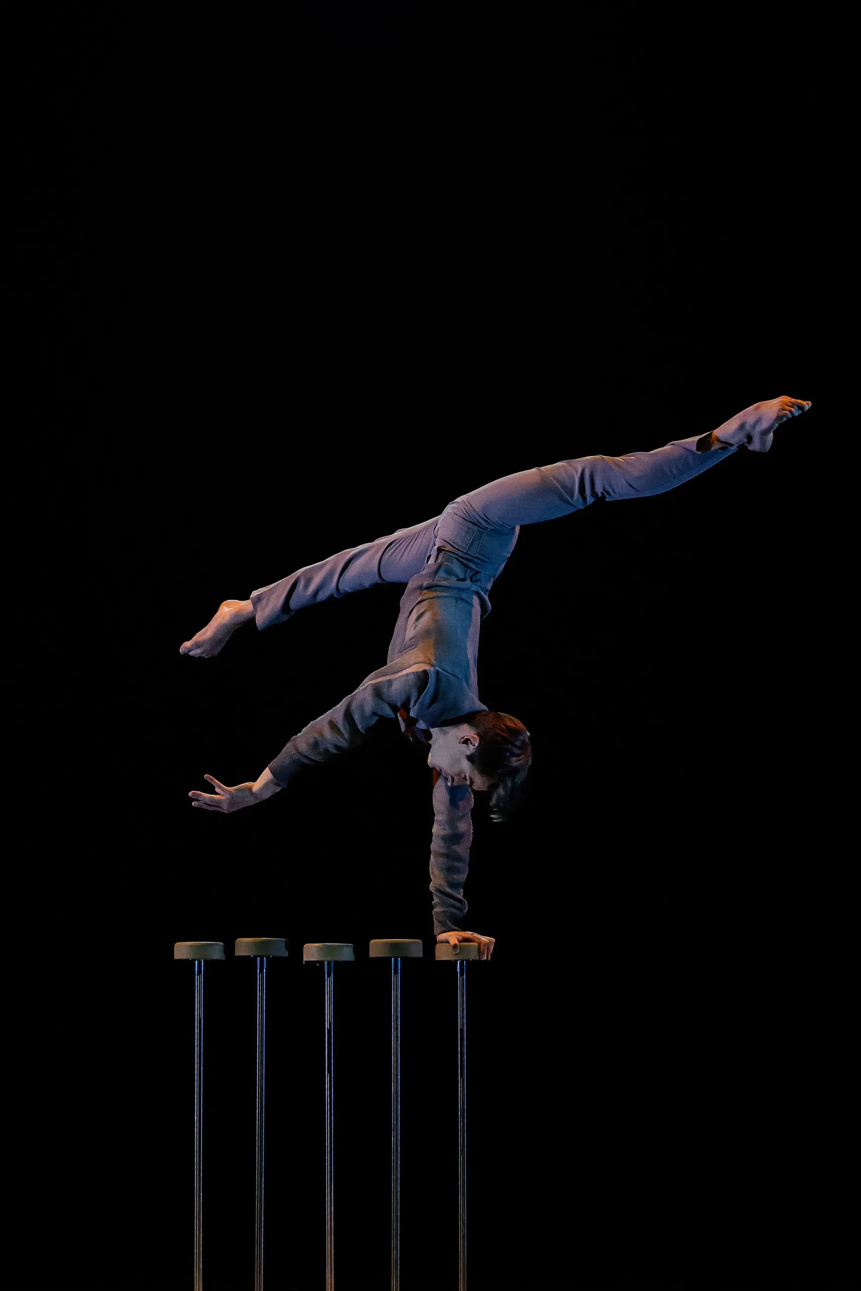 Louis-Thomas Gauthier, vêtu d'une tenue grise, se tient en équilibre sur une main au sommet de quatre grands supports sur un fond sombre, les jambes tendues en fente, faisant preuve d'une force et d'une souplesse remarquables.