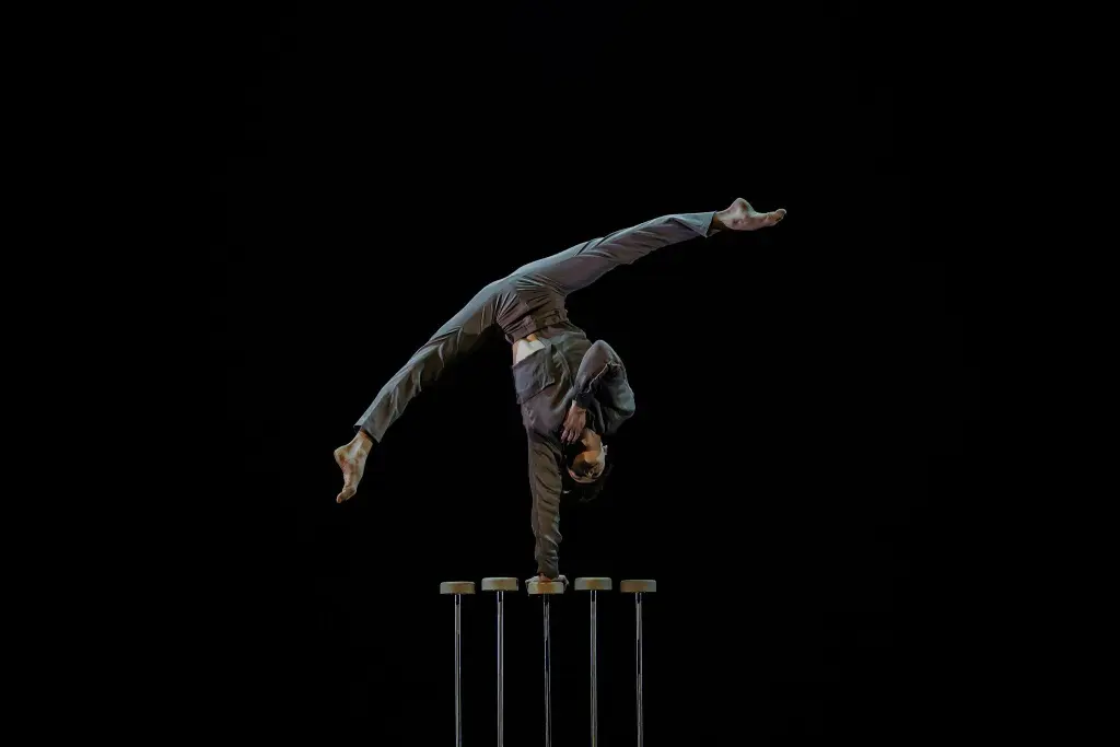 Une personne en costume, évoquant Louis-Thomas Gauthier, effectue un équilibre à une main sur de grandes cannes d'équilibre sur fond noir, les jambes étendues dans un élégant dédoublement.