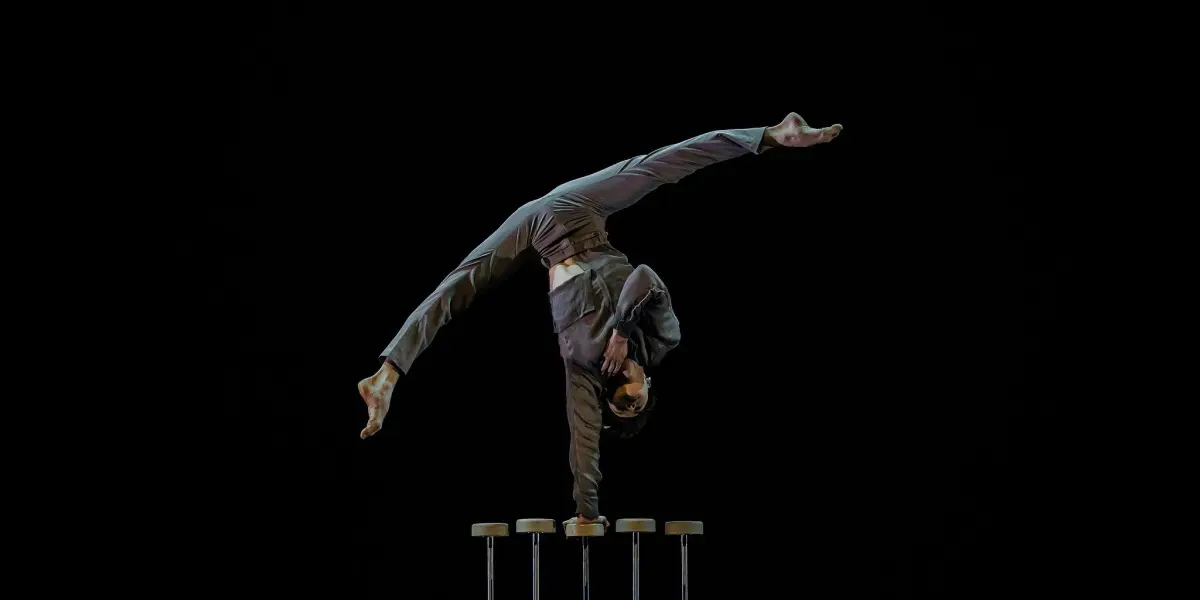 Une personne en costume, évoquant Louis-Thomas Gauthier, effectue un équilibre à une main sur de grandes cannes d'équilibre sur fond noir, les jambes étendues dans un élégant dédoublement.