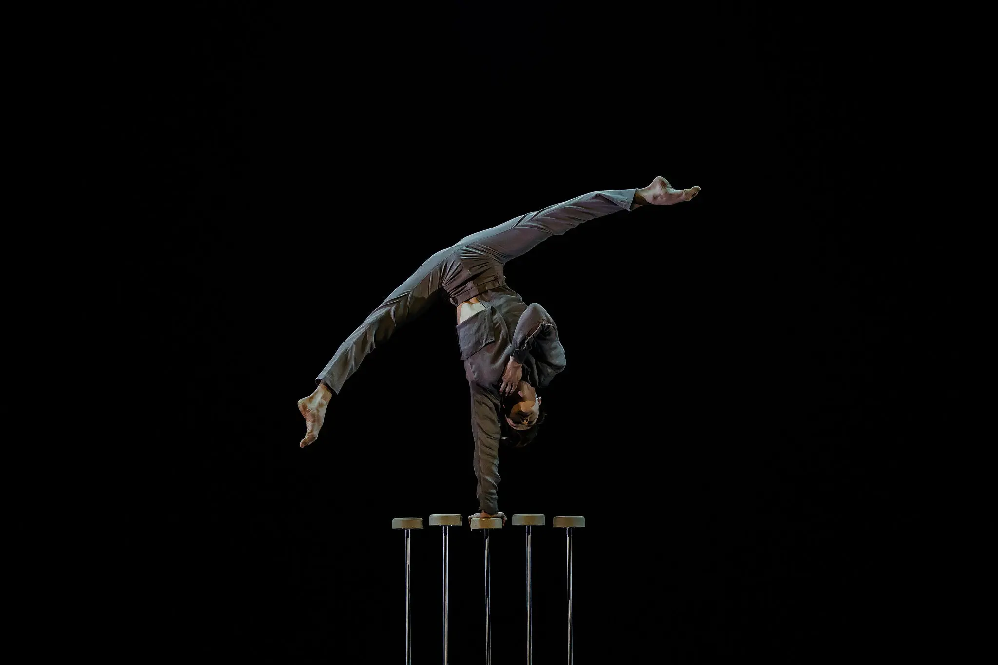 Une personne en costume, évoquant Louis-Thomas Gauthier, effectue un équilibre à une main sur de grandes cannes d'équilibre sur fond noir, les jambes étendues dans un élégant dédoublement.
