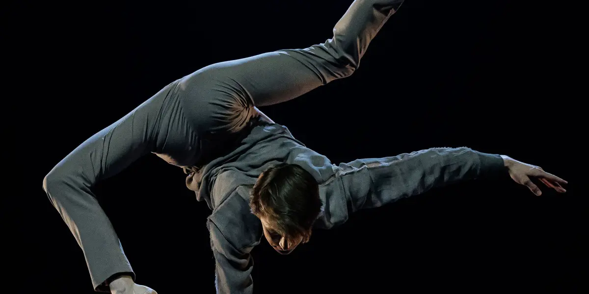 Louis-Thomas Gauthier, un artiste vêtu de gris, se tient en équilibre sur une main au sommet de poteaux parallèles, les jambes tendues en fente, faisant preuve de force et de souplesse sur un fond sombre.