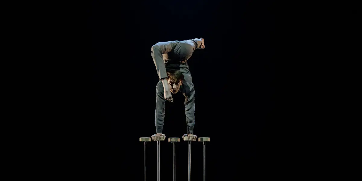 Louis-Thomas Gauthier fait l'équilibre sur quatre grands poteaux, en pliant son corps vers l'arrière avec une jambe tendue, sur un fond noir et sombre.