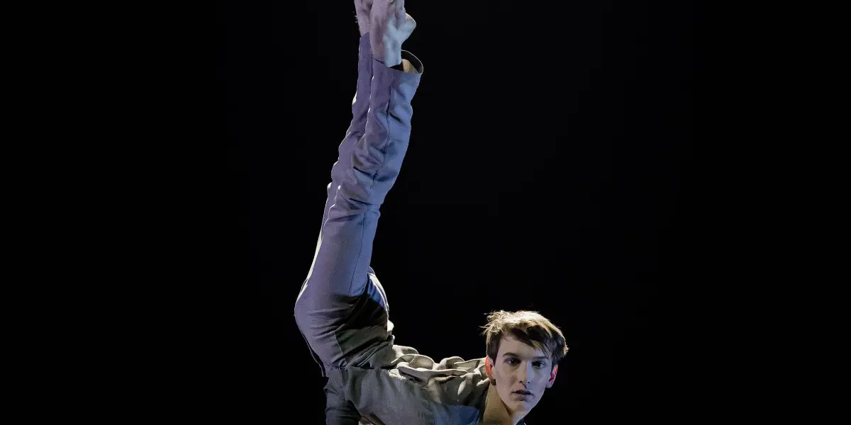 Louis-Thomas Gauthier, vêtu d'un costume gris, effectue un équilibre sur une main sur des barres parallèles - une jambe pointée vers le haut, l'autre pliée - sur un fond sombre et sous un projecteur.