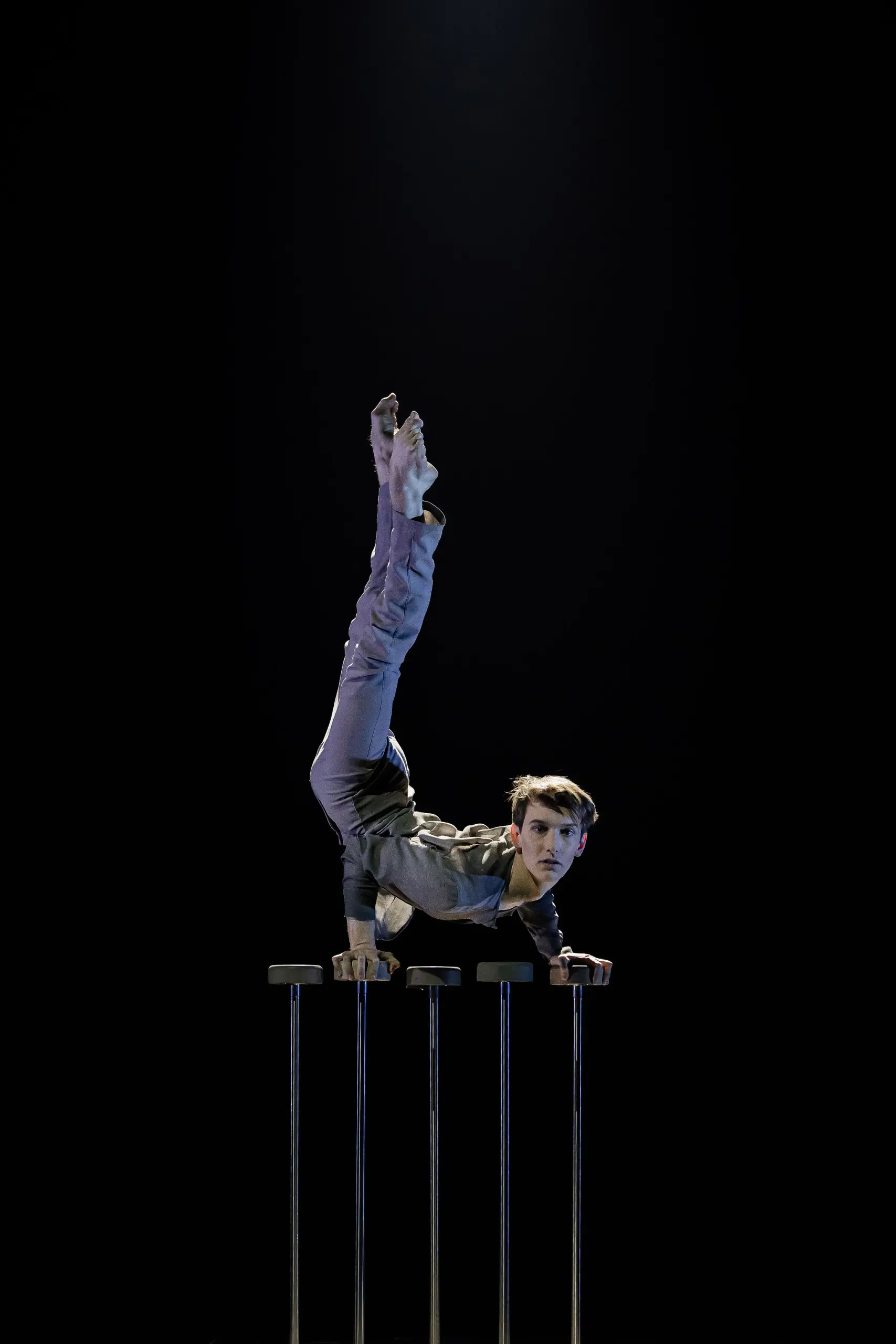 Louis-Thomas Gauthier, vêtu d'un costume gris, effectue un équilibre sur une main sur des barres parallèles - une jambe pointée vers le haut, l'autre pliée - sur un fond sombre et sous un projecteur.