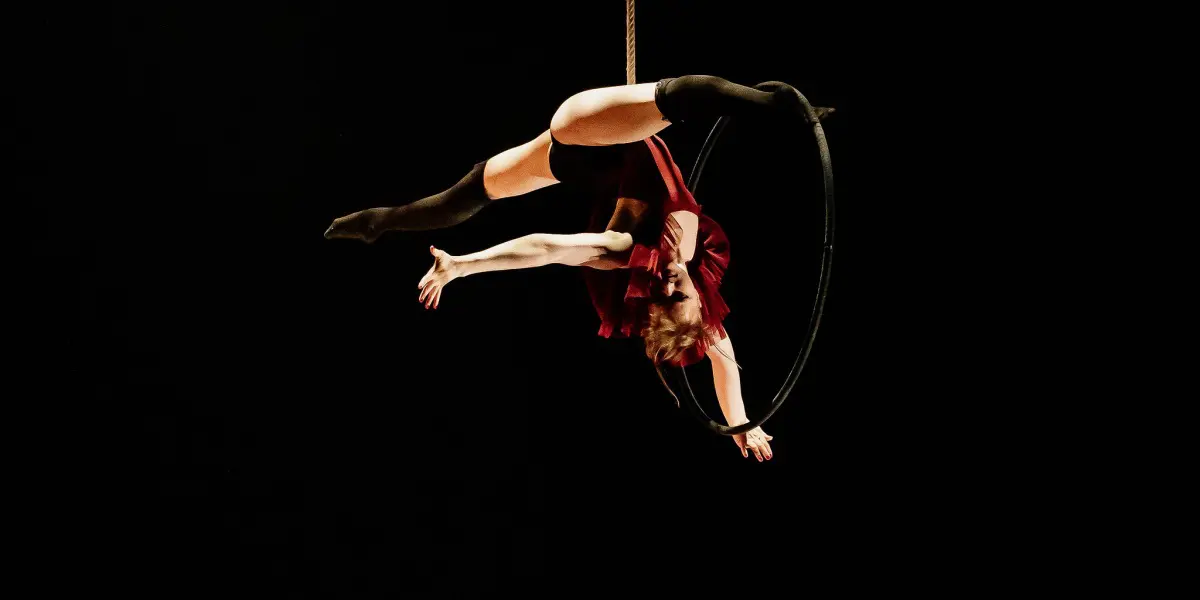 L'artiste aérienne Esmé Higgon, vêtue d'un costume rouge, est suspendue à l'envers à un cerceau sur un fond sombre, les bras et les jambes gracieusement étendus.