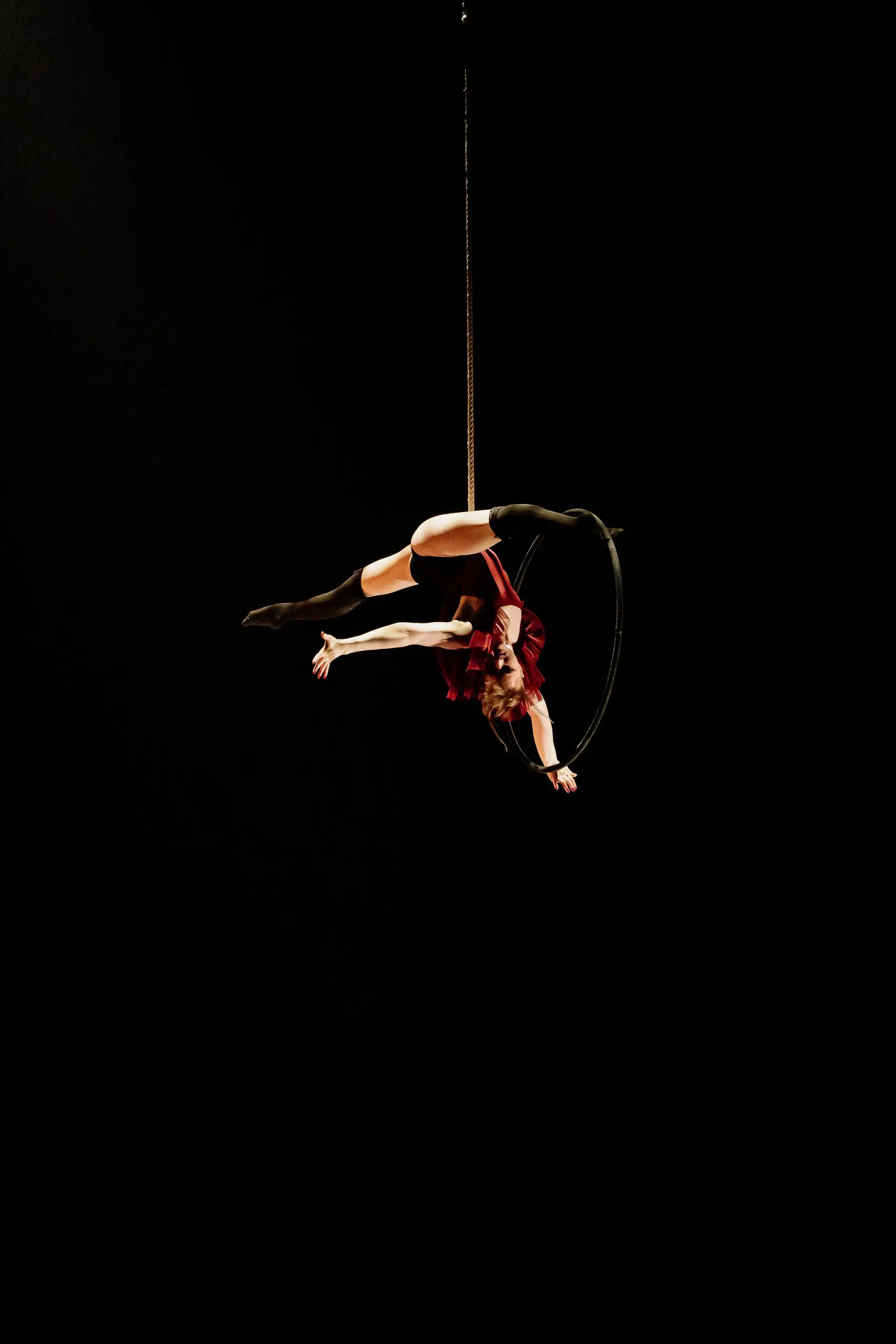 L'artiste aérienne Esmé Higgon, vêtue d'un costume rouge, est suspendue à l'envers à un cerceau sur un fond sombre, les bras et les jambes gracieusement étendus.