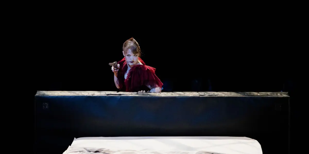 Esmé Higgon, vêtue d'une robe rouge spectaculaire, se tient derrière une grande structure noire, tenant un pistolet et regardant attentivement vers l'avant. Au premier plan, un lit défait avec des draps blancs est visible sur l'arrière-plan sombre et minimal.