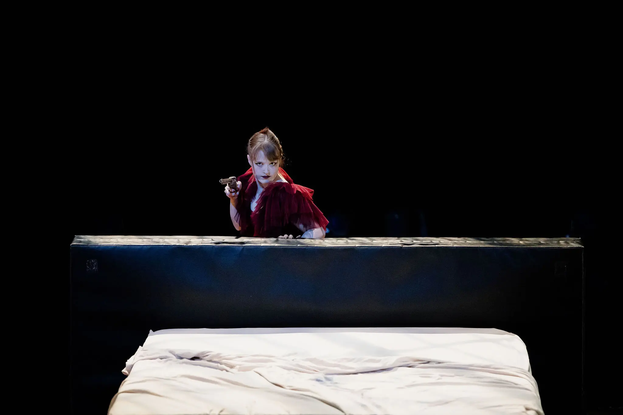 Esmé Higgon, vêtue d'une robe rouge spectaculaire, se tient derrière une grande structure noire, tenant un pistolet et regardant attentivement vers l'avant. Au premier plan, un lit défait avec des draps blancs est visible sur l'arrière-plan sombre et minimal.
