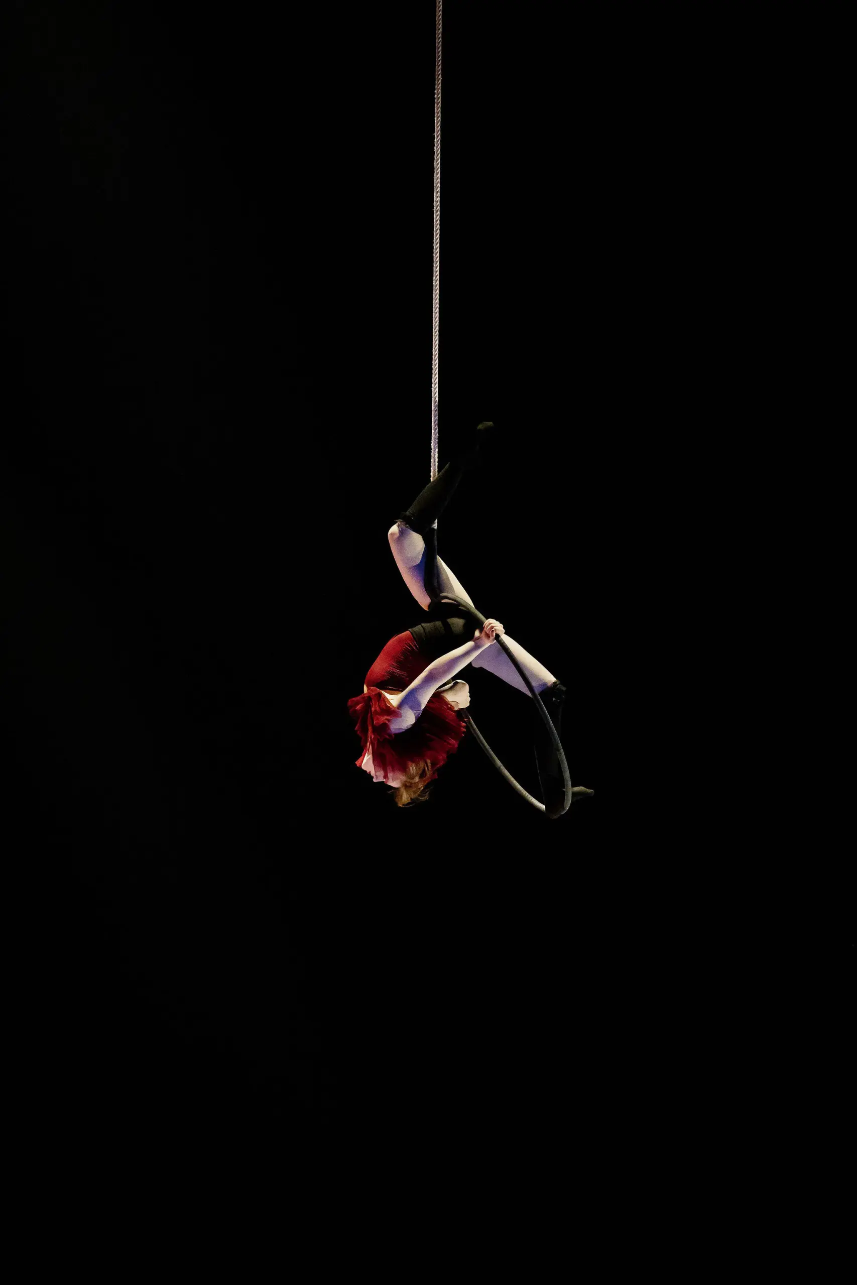 L'artiste aérienne Esmé Higgon, vêtue d'un costume rouge, est suspendue à l'envers à un cerceau sur un fond noir, se tenant au cerceau avec ses jambes et s'agrippant à la corde au-dessus.