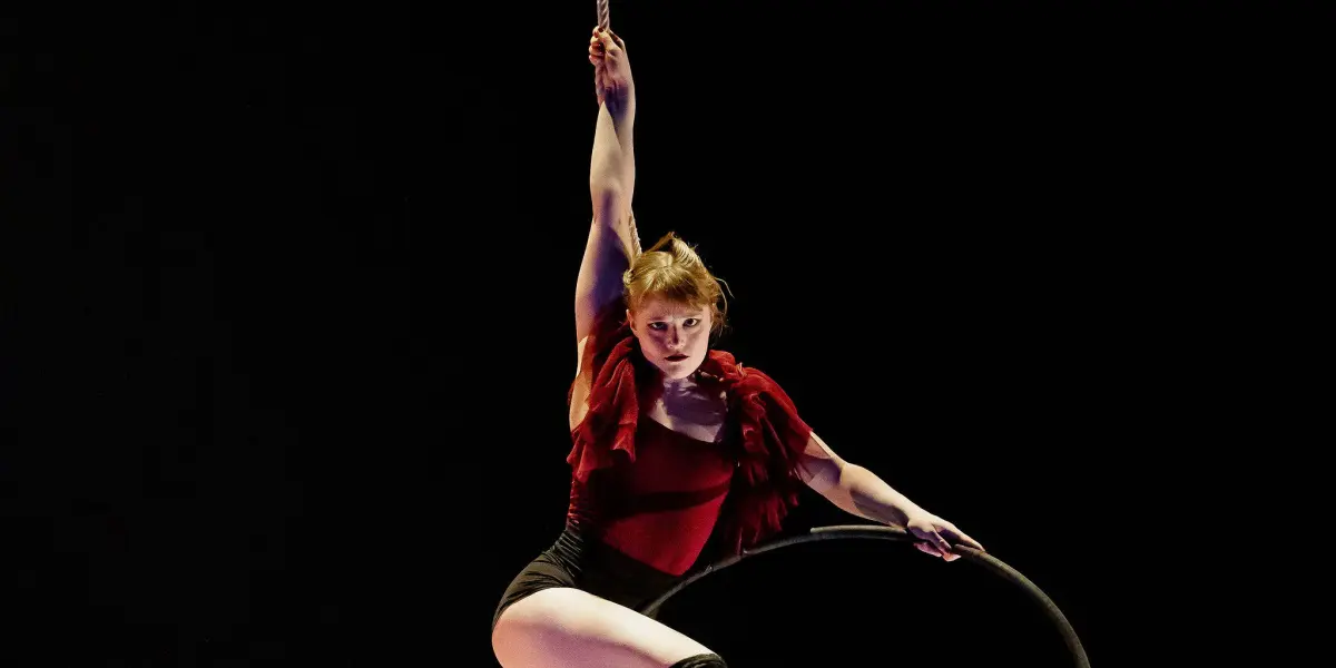 Une acrobate, Esmé Higgon, vêtue d'un haut rouge et d'un short noir, s'exécute sur un cerceau aérien suspendu, saisissant la corde d'une main et posant gracieusement sur un fond sombre.