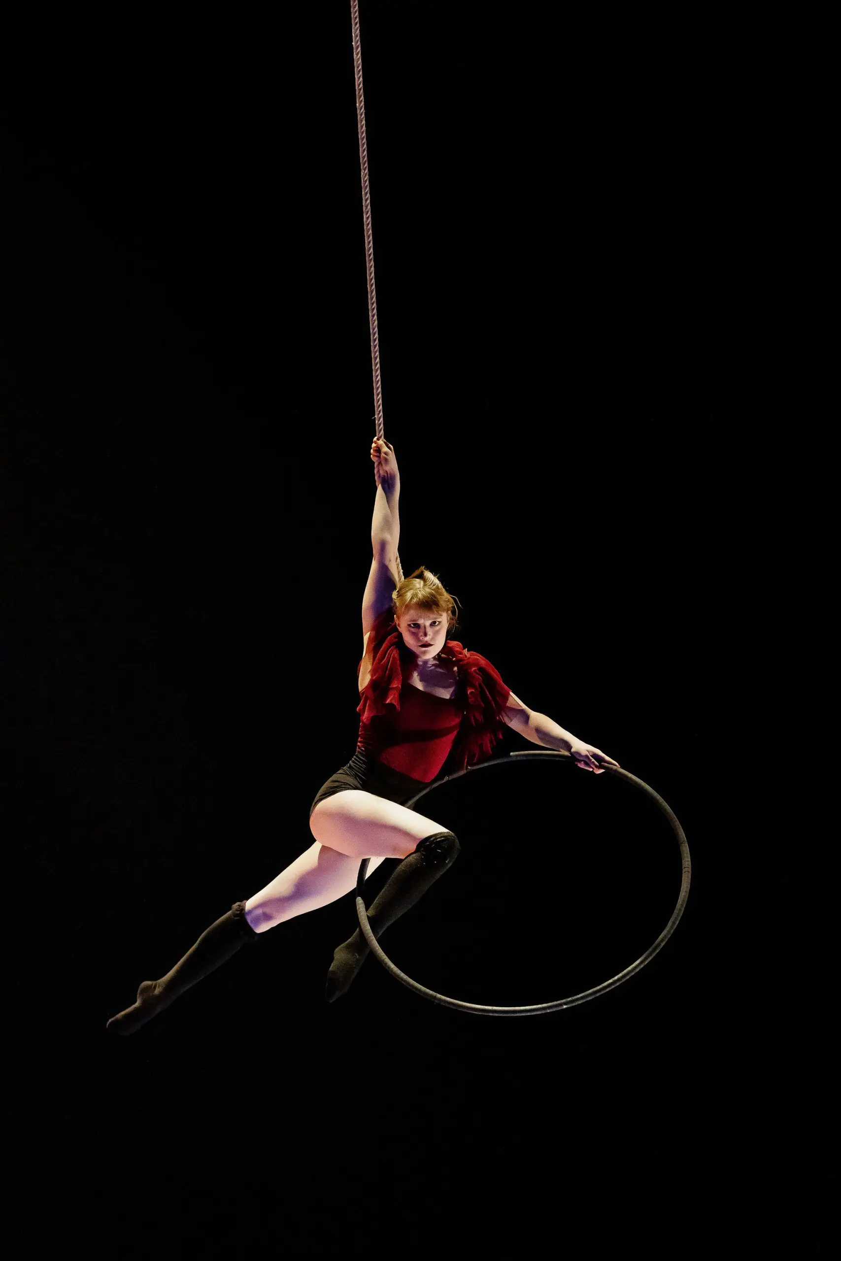 Une acrobate, Esmé Higgon, vêtue d'un haut rouge et d'un short noir, s'exécute sur un cerceau aérien suspendu, saisissant la corde d'une main et posant gracieusement sur un fond sombre.