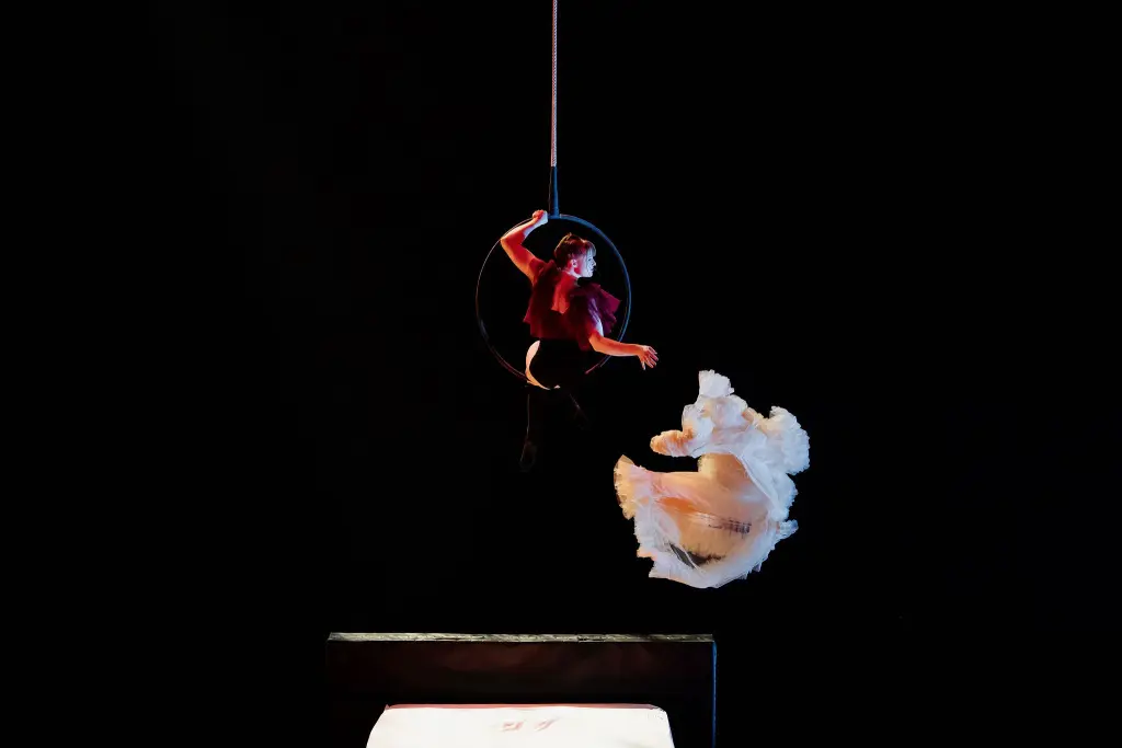 Une artiste vêtue d'un haut rouge est suspendue à un cerceau aérien au-dessus d'un lit, tandis qu'Esmé Higgon, vêtue d'une robe blanche fluide, saute de façon spectaculaire à côté d'elles sur un fond sombre.