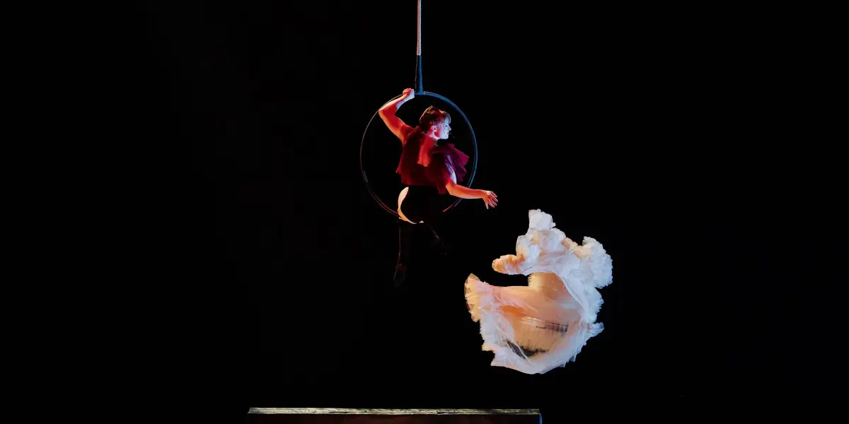 Une artiste vêtue d'un haut rouge est suspendue à un cerceau aérien au-dessus d'un lit, tandis qu'Esmé Higgon, vêtue d'une robe blanche fluide, saute de façon spectaculaire à côté d'elles sur un fond sombre.