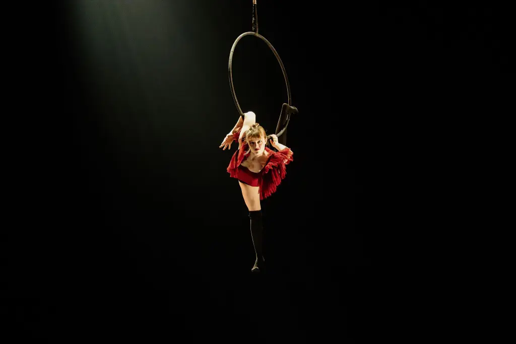 Une voltigeuse, Esmé Higgon, vêtue d'une tenue rouge, exécute une fente tout en étant suspendue à un cerceau sur un fond sombre, un projecteur mettant en valeur sa souplesse et sa force.