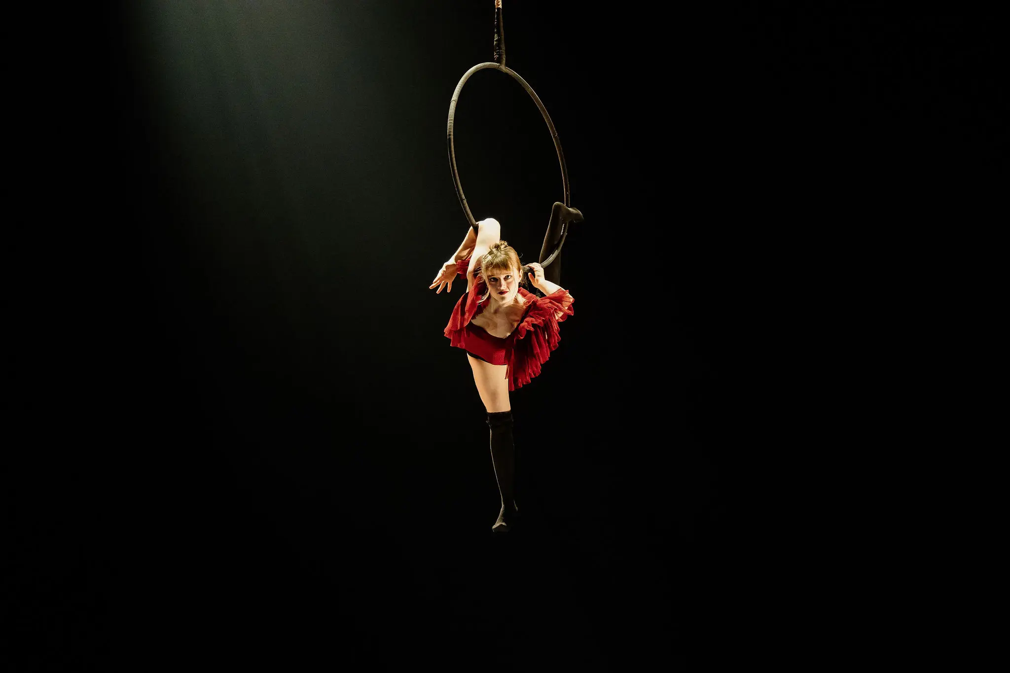 Une voltigeuse, Esmé Higgon, vêtue d'une tenue rouge, exécute une fente tout en étant suspendue à un cerceau sur un fond sombre, un projecteur mettant en valeur sa souplesse et sa force.