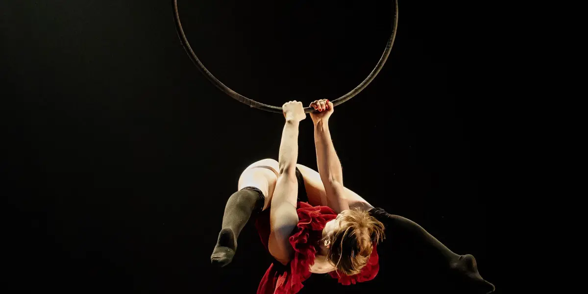 L'artiste aérienne Esmé Higgon, vêtue d'un costume rouge, se produit sur un cerceau suspendu sur un fond sombre, suspendue à l'envers, les jambes étendues, et saisissant le cerceau à deux mains.