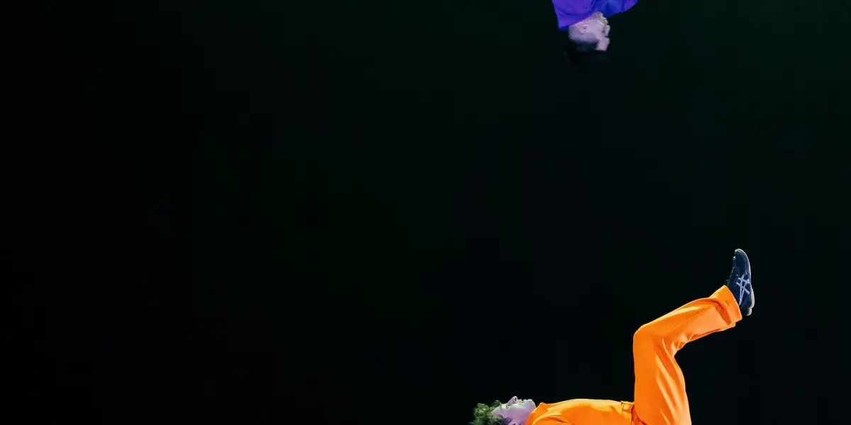 Deux acrobates aux tenues colorées se produisent sur scène ; l'un, en orange, se tient en équilibre sur le dos sur une plate-forme, tandis que l'autre - rappelant le style audacieux de Victor Crepin - est en train de faire un saut en hauteur, sur un fond sombre parsemé de blocs vibrants.