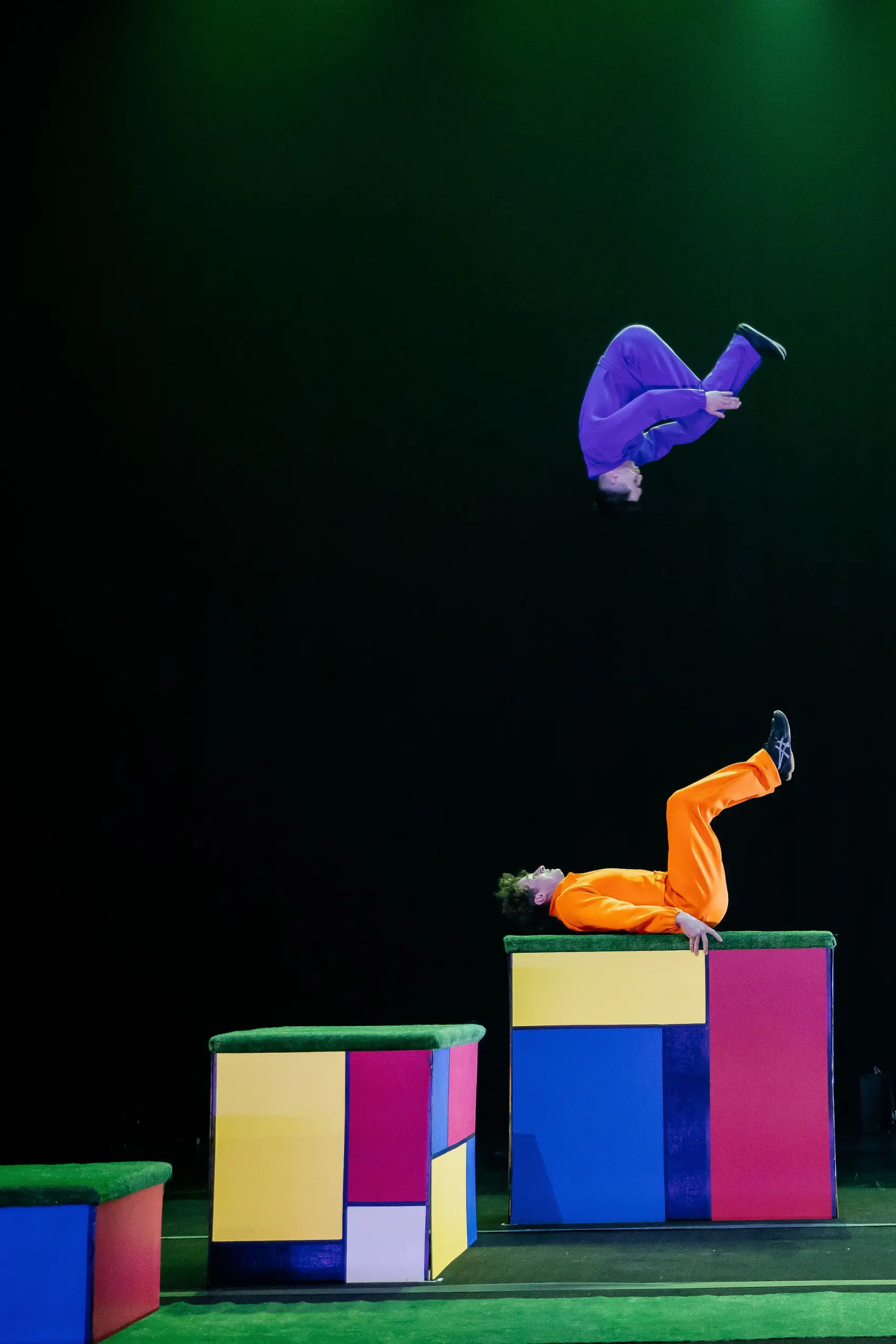 Deux acrobates aux tenues colorées se produisent sur scène ; l'un, en orange, se tient en équilibre sur le dos sur une plate-forme, tandis que l'autre - rappelant le style audacieux de Victor Crepin - est en train de faire un saut en hauteur, sur un fond sombre parsemé de blocs vibrants.