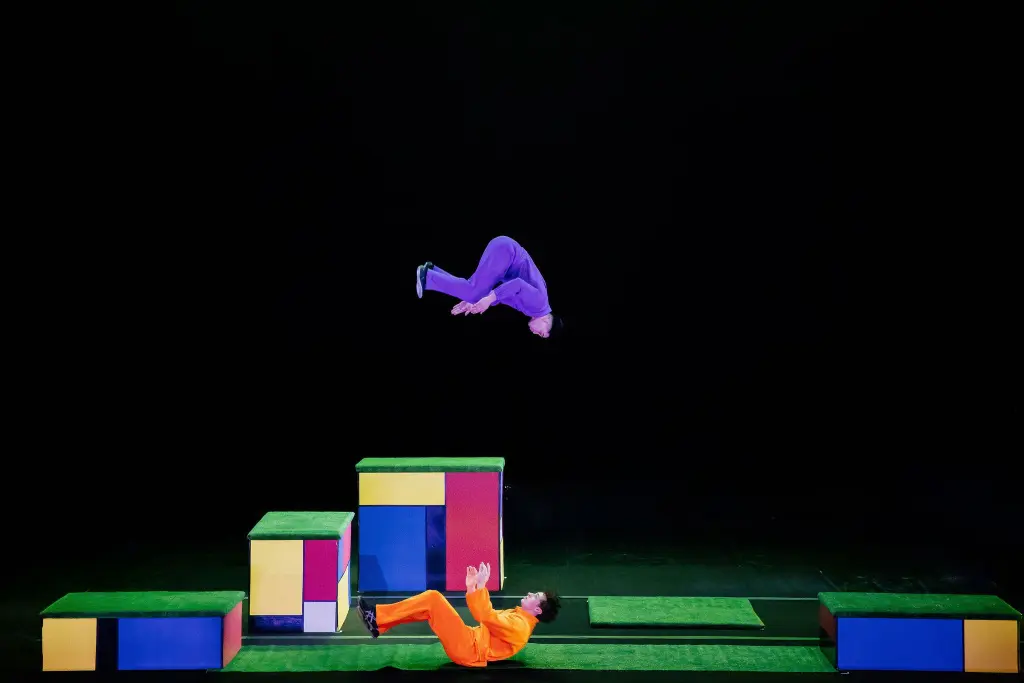 Un artiste en tenue violette fait un saut périlleux arrière au-dessus de plates-formes de blocs colorés, tandis que Victor Crepin, habillé en orange, est allongé sur le dos sur la scène en dessous, en tendant les mains vers le haut. L'arrière-plan est noir.
