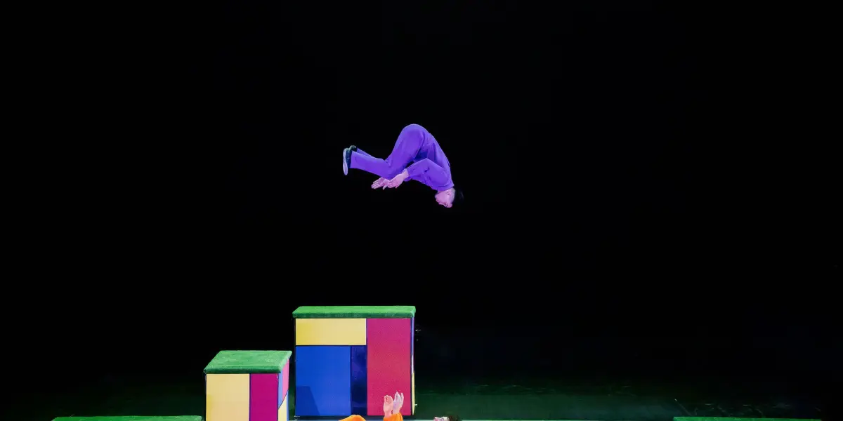 Un artiste en tenue violette fait un saut périlleux arrière au-dessus de plates-formes de blocs colorés, tandis que Victor Crepin, habillé en orange, est allongé sur le dos sur la scène en dessous, en tendant les mains vers le haut. L'arrière-plan est noir.