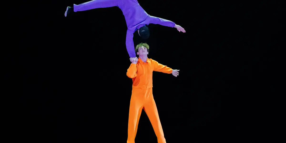 Deux acrobates réalisent un numéro d'équilibre sur des blocs colorés. L'un, en tenue orange, se tient sur les blocs tout en soutenant Victor Crépin, en tenue violette, qui est à l'envers, une main sur la tête de l'autre.