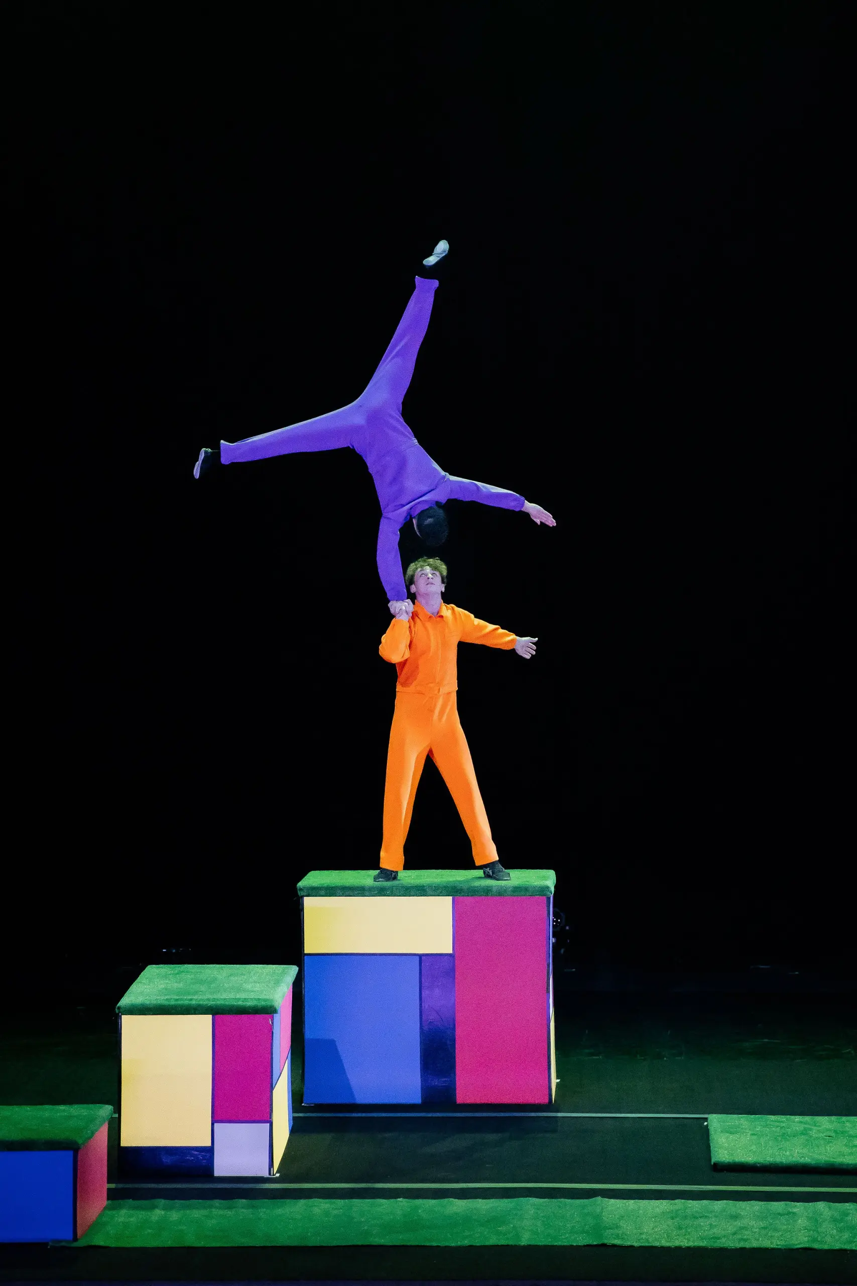 Deux acrobates réalisent un numéro d'équilibre sur des blocs colorés. L'un, en tenue orange, se tient sur les blocs tout en soutenant Victor Crépin, en tenue violette, qui est à l'envers, une main sur la tête de l'autre.