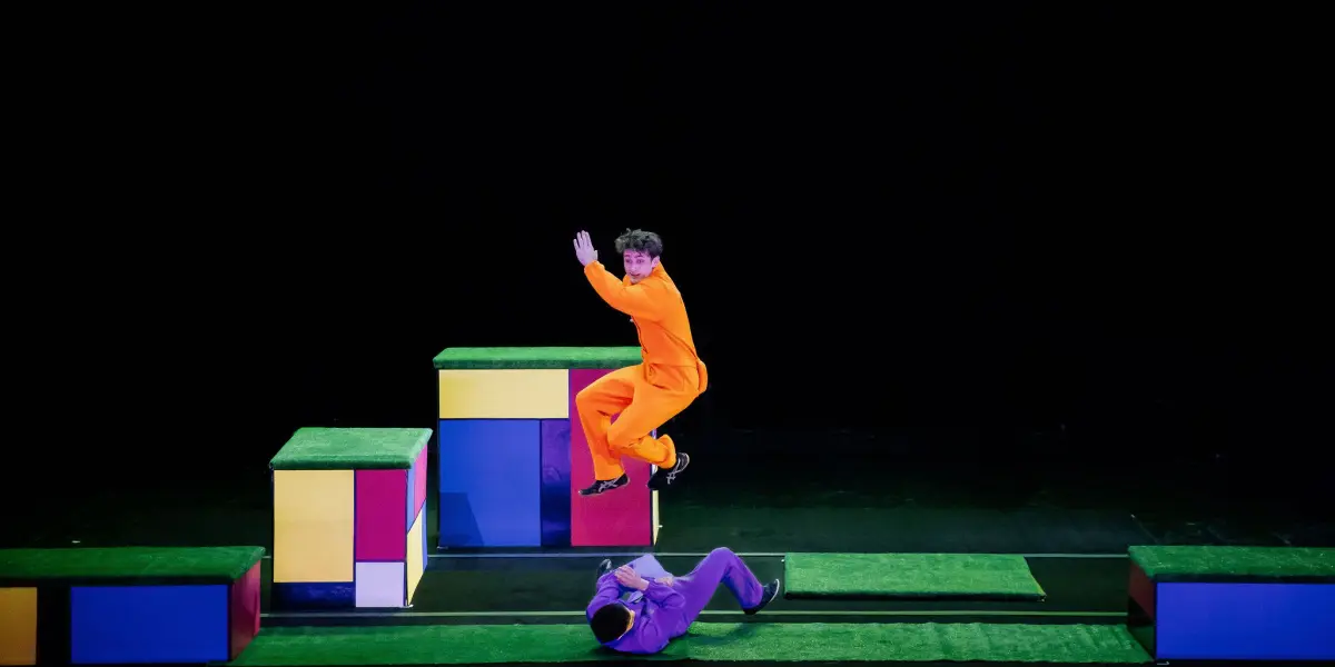 Un artiste vêtu d'une combinaison orange saute en l'air tandis qu'un autre, peut-être Victor Crepin, est allongé sur le sol. Ils se trouvent sur une scène composée de blocs géométriques colorés et de plates-formes, sur un fond noir.