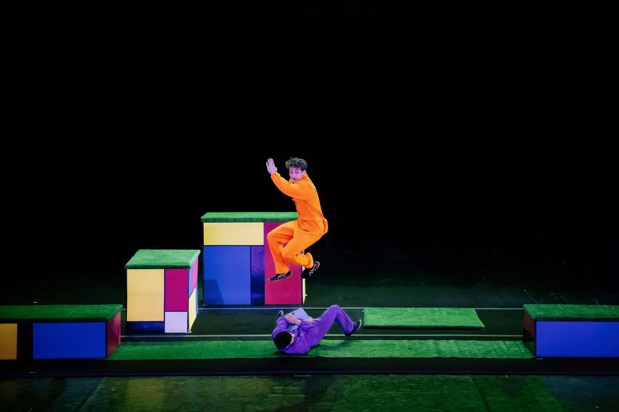 Un artiste vêtu d'une combinaison orange saute en l'air tandis qu'un autre, peut-être Victor Crepin, est allongé sur le sol. Ils se trouvent sur une scène composée de blocs géométriques colorés et de plates-formes, sur un fond noir.