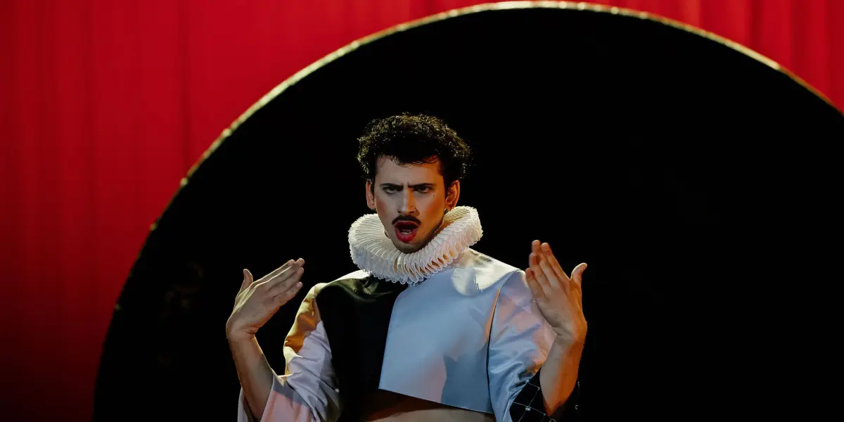 Un artiste en costume dramatique avec un col à volants blanc et une tenue géométrique fait des gestes expressifs sur scène, devant un rideau rouge et une grande toile de fond circulaire noire dans le style de Joaquin Barral.