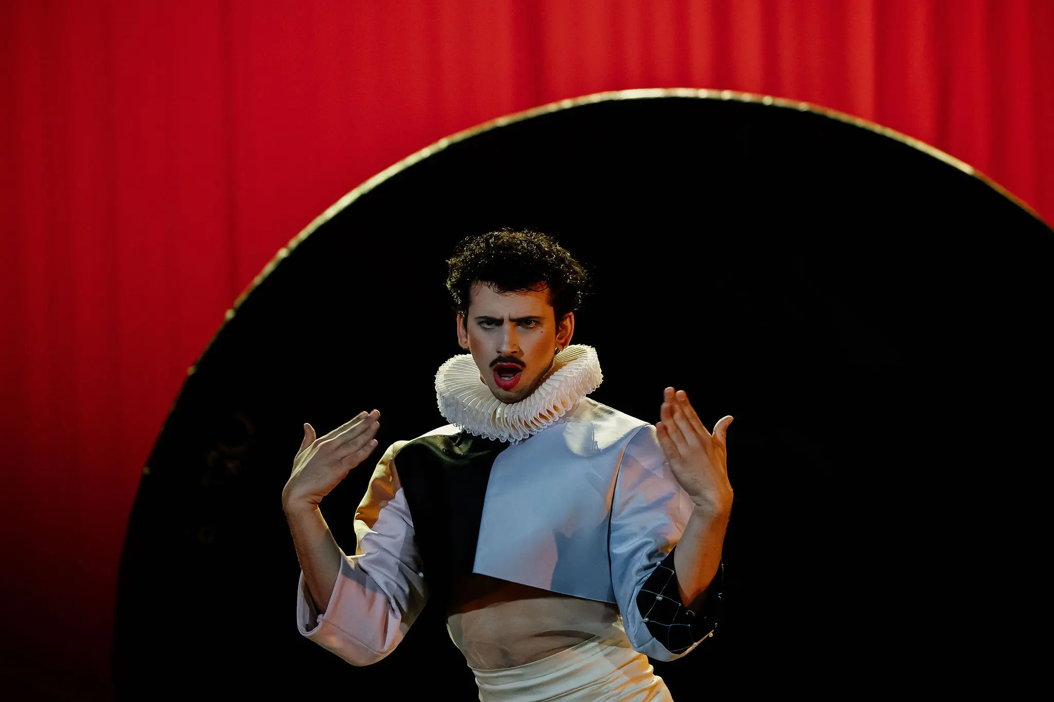 Un artiste en costume dramatique avec un col à volants blanc et une tenue géométrique fait des gestes expressifs sur scène, devant un rideau rouge et une grande toile de fond circulaire noire dans le style de Joaquin Barral.