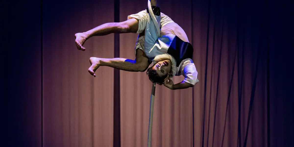 L'artiste aérien Joaquin Barral, vêtu de vêtements légers, est suspendu à l'envers sur une corde verticale, les jambes accrochées, avec un éclairage de scène dramatique et un rideau sombre en toile de fond.