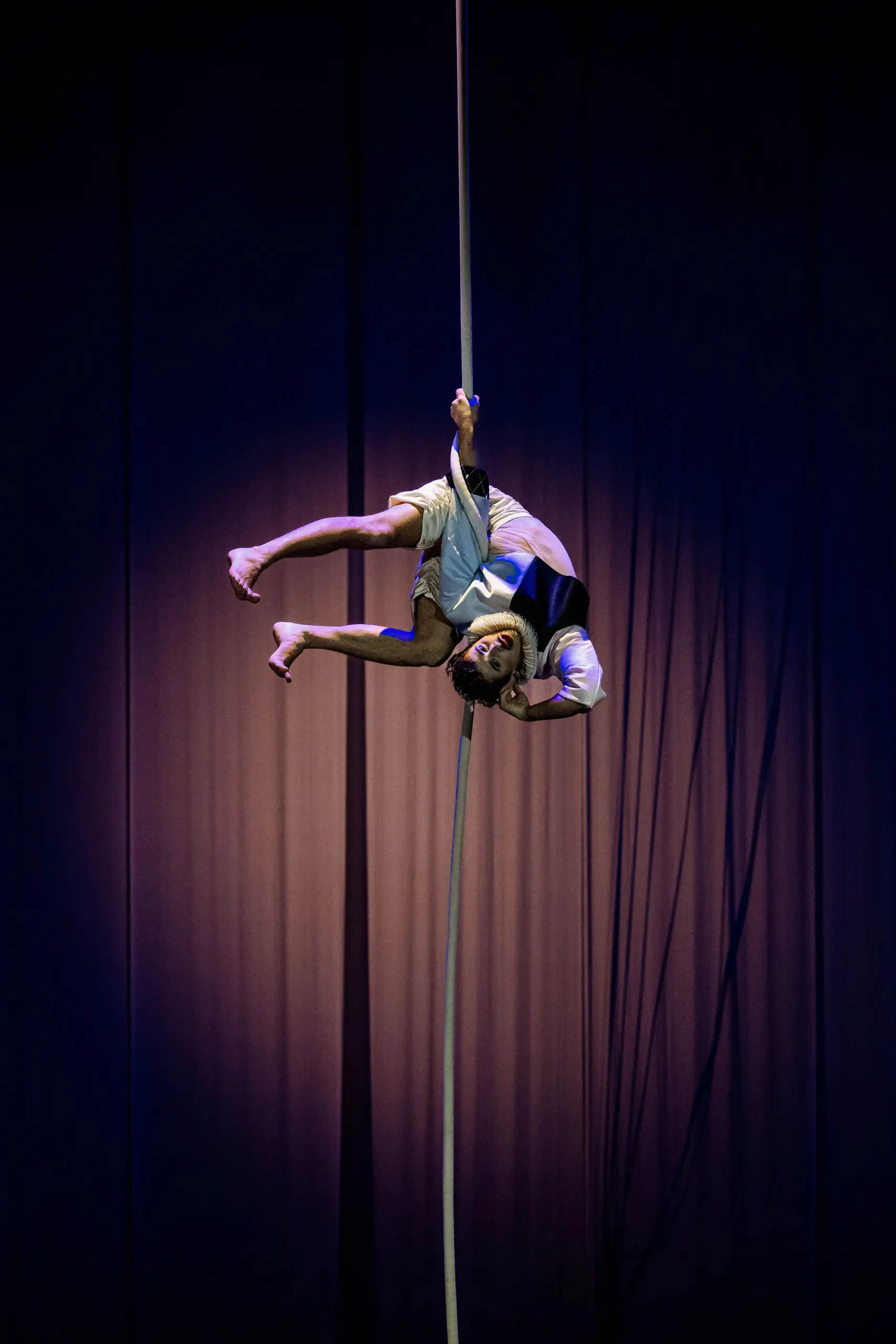 L'artiste aérien Joaquin Barral, vêtu de vêtements légers, est suspendu à l'envers sur une corde verticale, les jambes accrochées, avec un éclairage de scène dramatique et un rideau sombre en toile de fond.