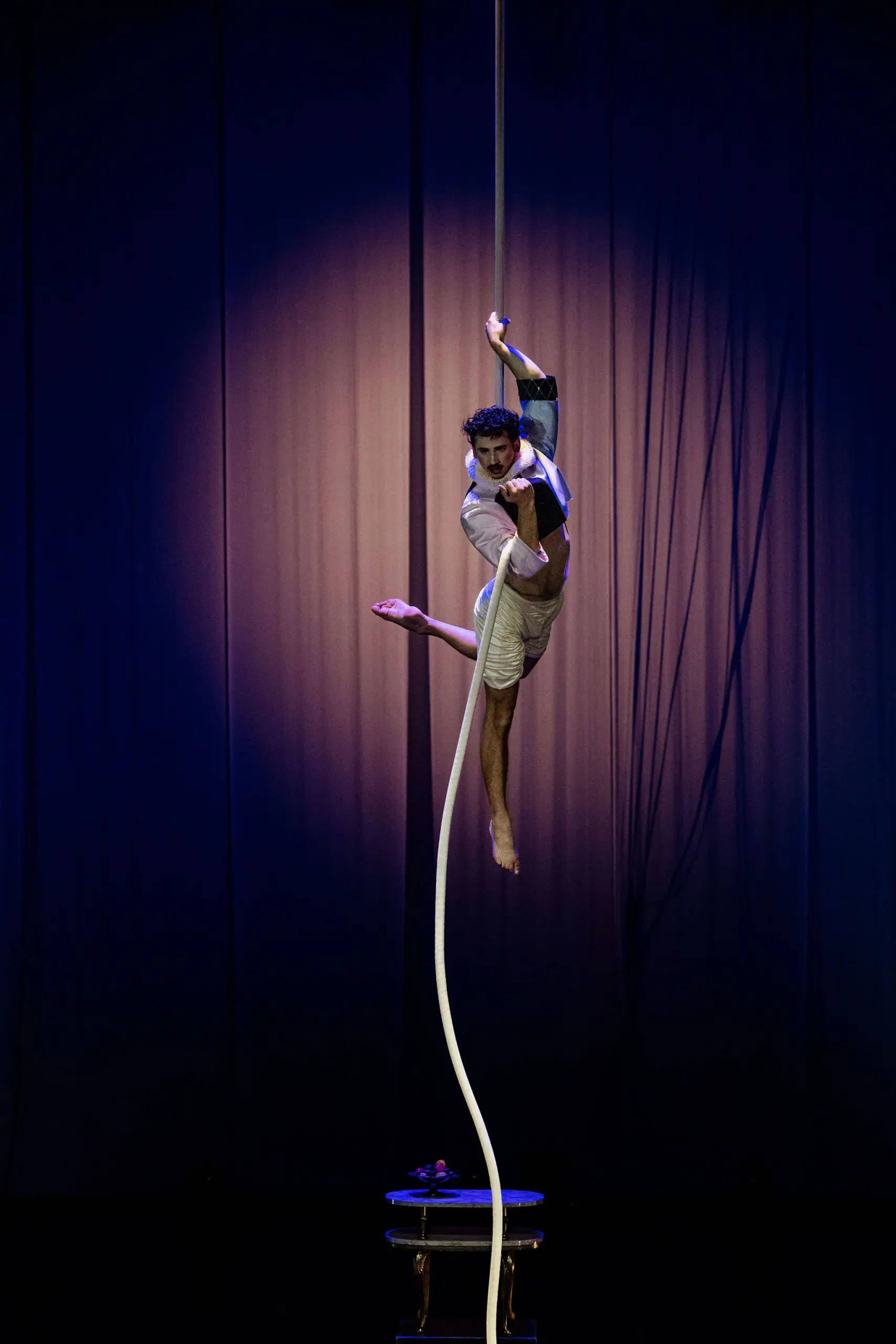 Un artiste aérien, Joaquin Barral, vêtu de blanc, se suspend gracieusement à une longue corde blanche sur une scène faiblement éclairée par un projecteur et un rideau violet en arrière-plan.