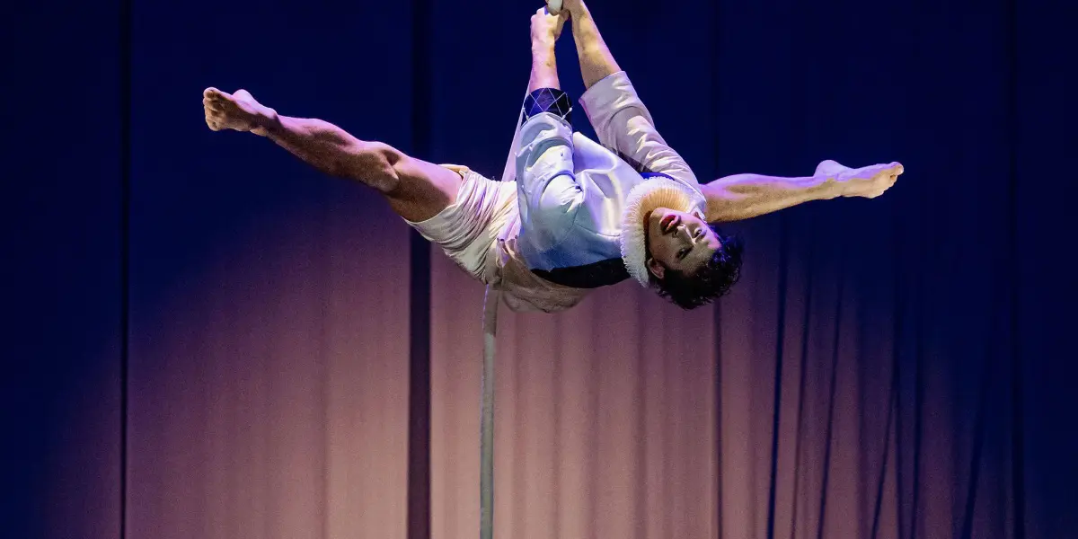 Un artiste de cirque, Joaquin Barral, en short et chemise blanche, est suspendu à l'envers à une corde verticale, qu'il saisit à deux mains tout en étendant ses jambes en éventail, sur un fond sombre éclairé en violet.