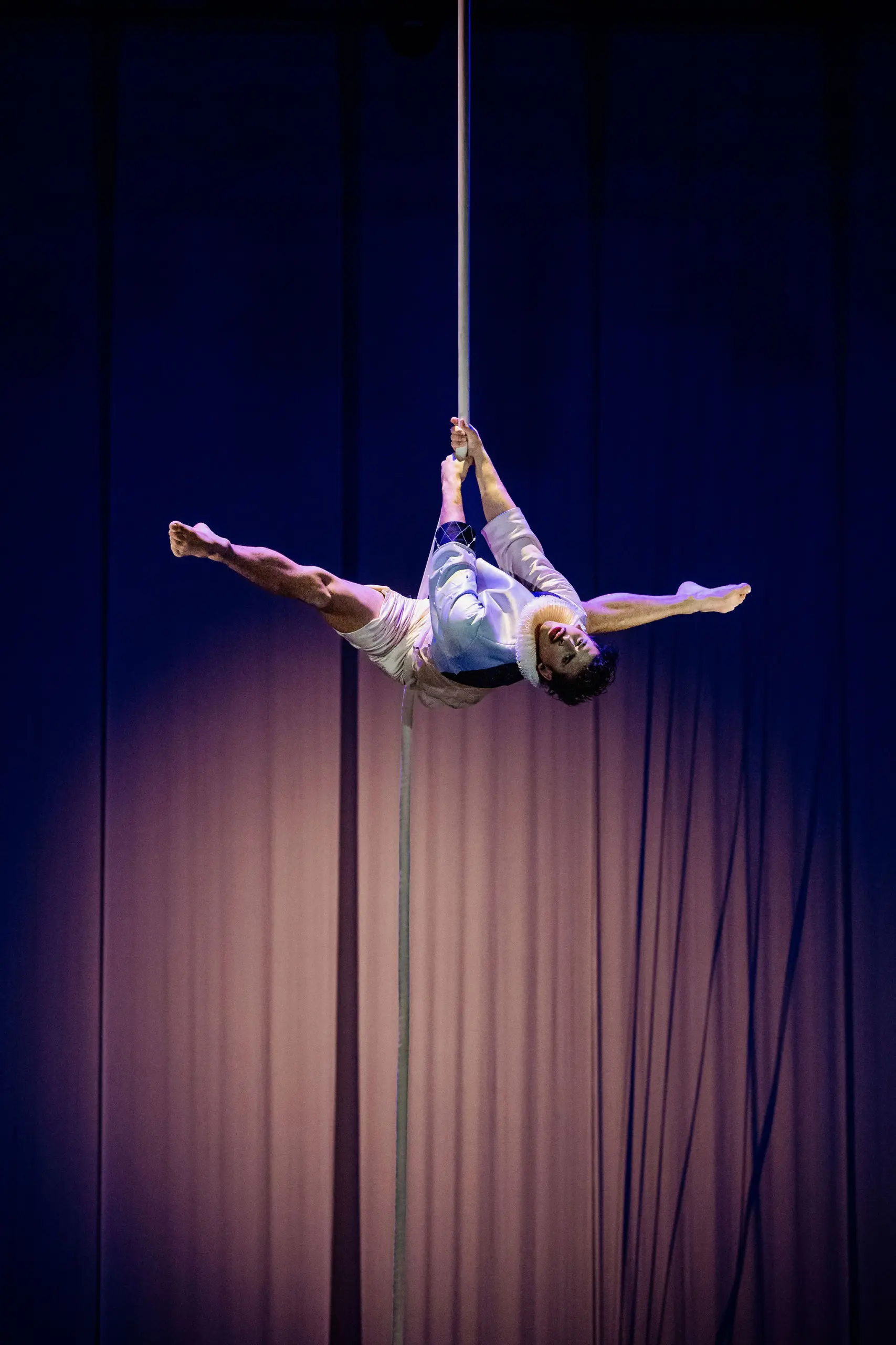 Un artiste de cirque, Joaquin Barral, en short et chemise blanche, est suspendu à l'envers à une corde verticale, qu'il saisit à deux mains tout en étendant ses jambes en éventail, sur un fond sombre éclairé en violet.