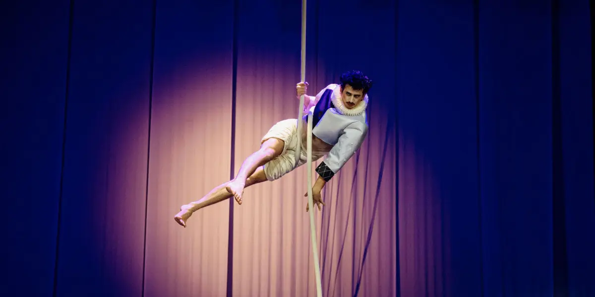 Un artiste vêtu d'une tenue légère et d'un col à volants pose dans les airs en se tenant à une corde verticale, avec un éclairage dramatique et des rideaux bleus en arrière-plan - une scène qui rappelle l'art captivant de Joaquin Barral.