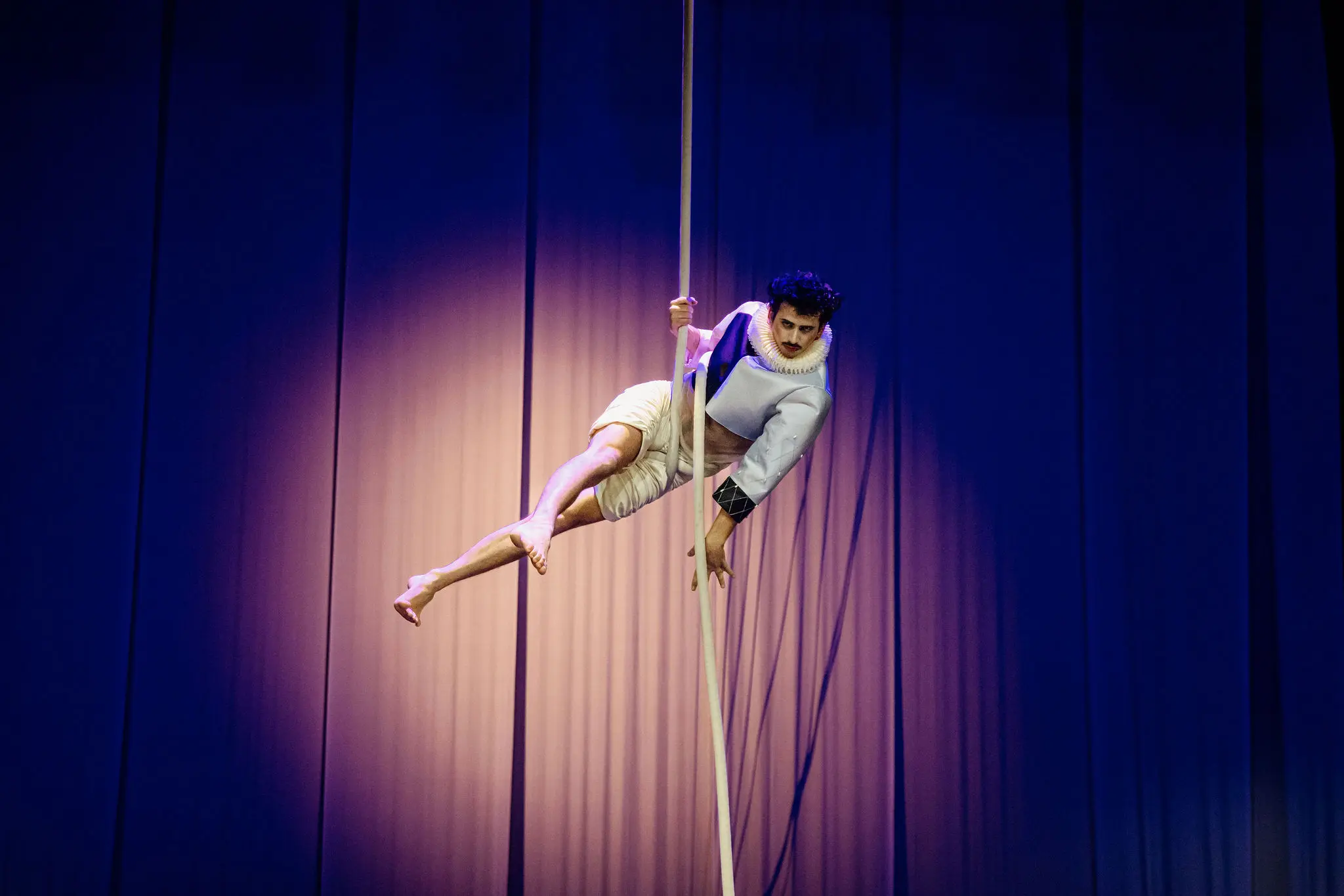 Un artiste vêtu d'une tenue légère et d'un col à volants pose dans les airs en se tenant à une corde verticale, avec un éclairage dramatique et des rideaux bleus en arrière-plan - une scène qui rappelle l'art captivant de Joaquin Barral.