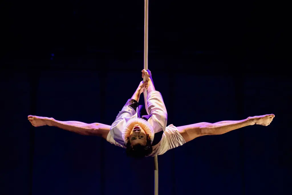 L'artiste aérien Joaquin Barral, vêtu d'un costume blanc, est suspendu à l'envers à un poteau vertical, qu'il saisit avec ses jambes, et étire ses bras et ses jambes dans un grand écart sur un fond sombre.