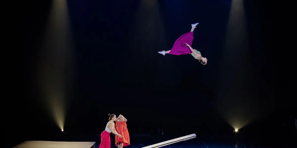 Une artiste en robe rose fait un saut périlleux au-dessus d'une bascule sur scène, tandis que Lilyclaire Major-Varallo et Marie-Sophie McKinnon en robes rouges se tiennent en dessous, sous les feux des projecteurs sur un fond sombre.