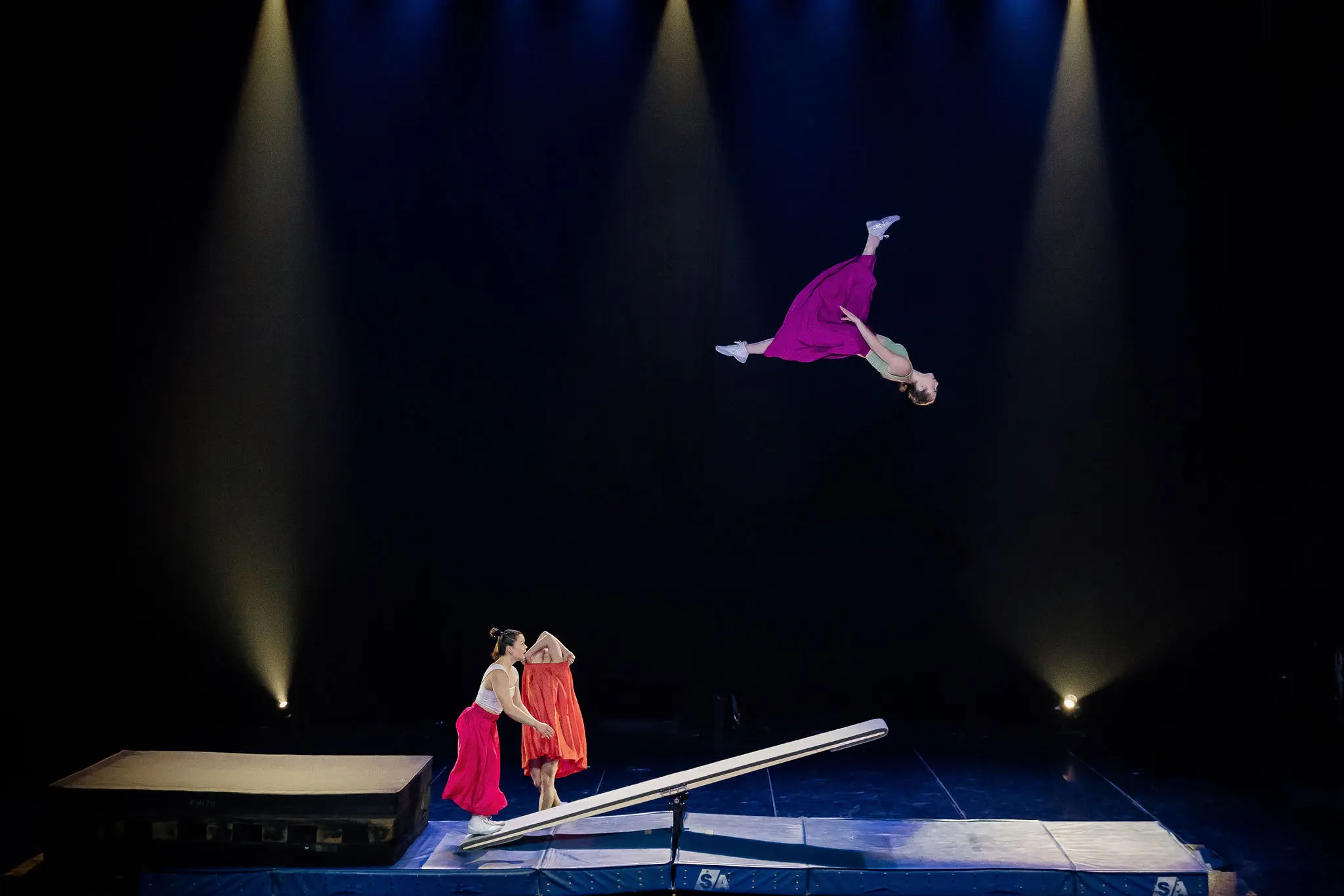 Une artiste en robe rose fait un saut périlleux au-dessus d'une bascule sur scène, tandis que Lilyclaire Major-Varallo et Marie-Sophie McKinnon en robes rouges se tiennent en dessous, sous les feux des projecteurs sur un fond sombre.