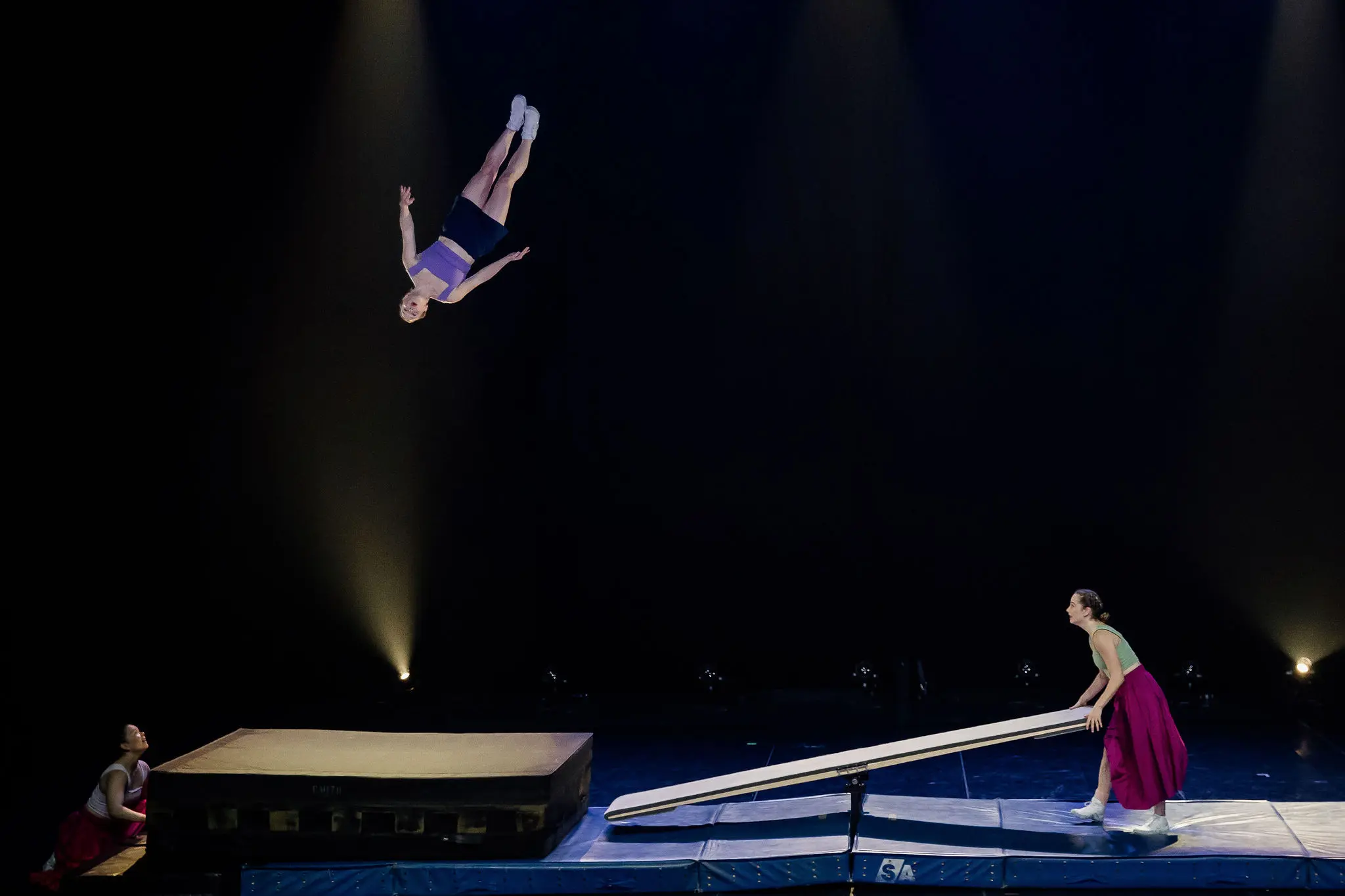Trois acrobates se produisent sur scène ; Lilyclaire Major-Varallo s'envole après avoir sauté d'une bascule, tandis qu'un autre manie la bascule et que Marie-Sophie McKinnon regarde à proximité. L'arrière-plan sombre met l'accent sur l'action.
