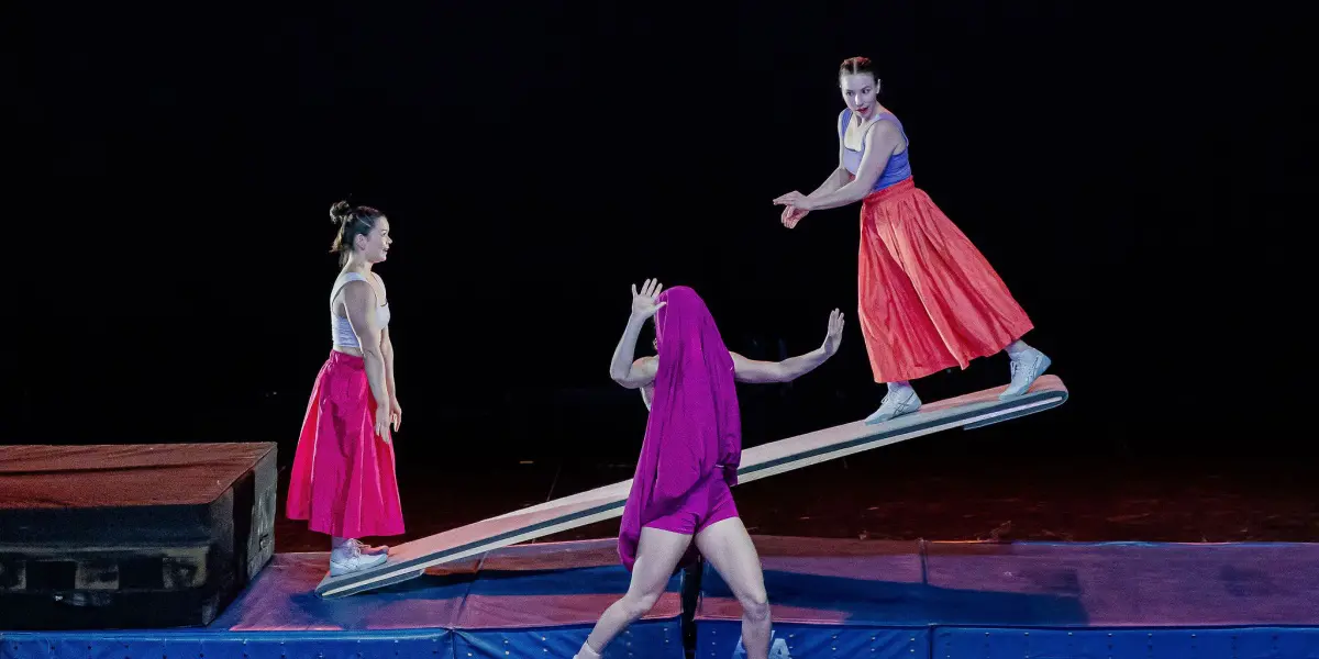 Trois artistes vêtus de tenues colorées se tiennent en équilibre sur une balançoire à bascule sur scène. Lilyclaire Major-Varallo se tient au sol avec un tissu sur la tête, tandis que Marie-Sophie McKinnon montre du doigt une extrémité de la balançoire et que la troisième interprète se tient en équilibre à l'opposé.